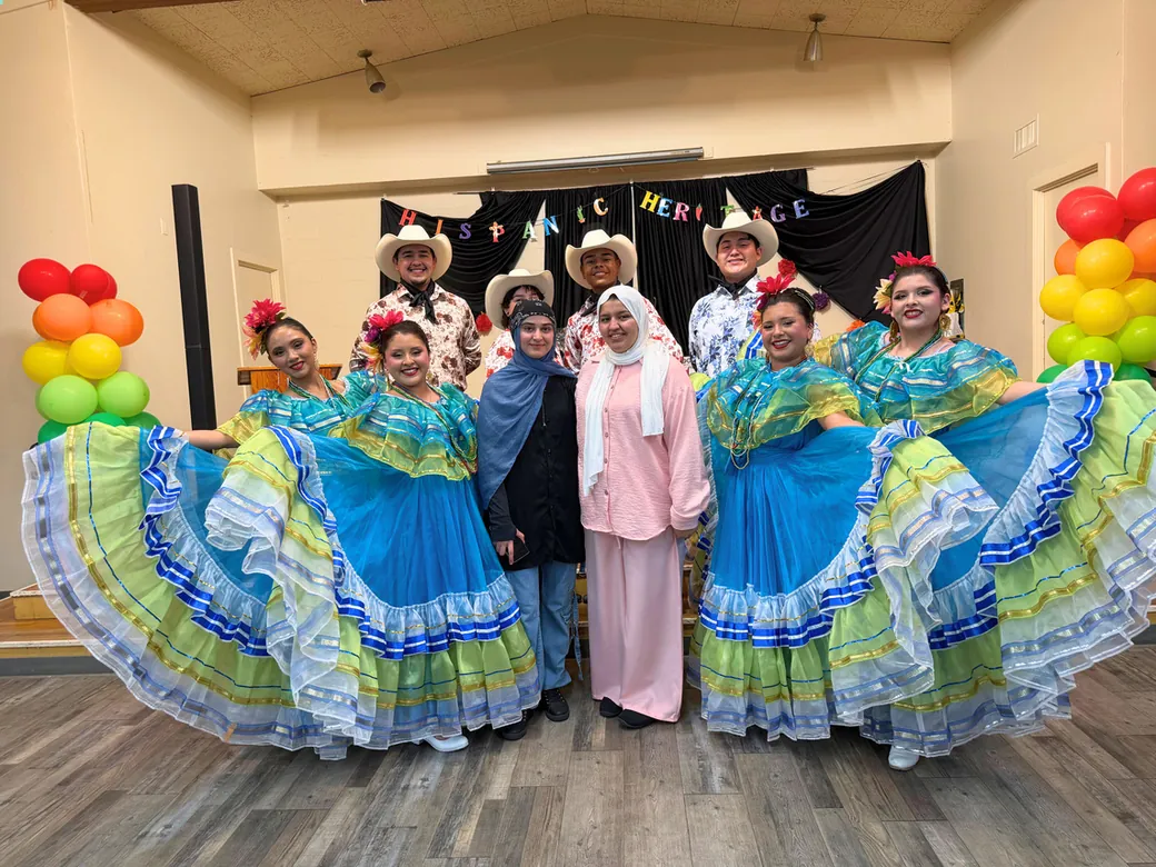 Folklorico dancers in green and blue dresses lined up under a balloon arch at a FIRM cultural celebration.