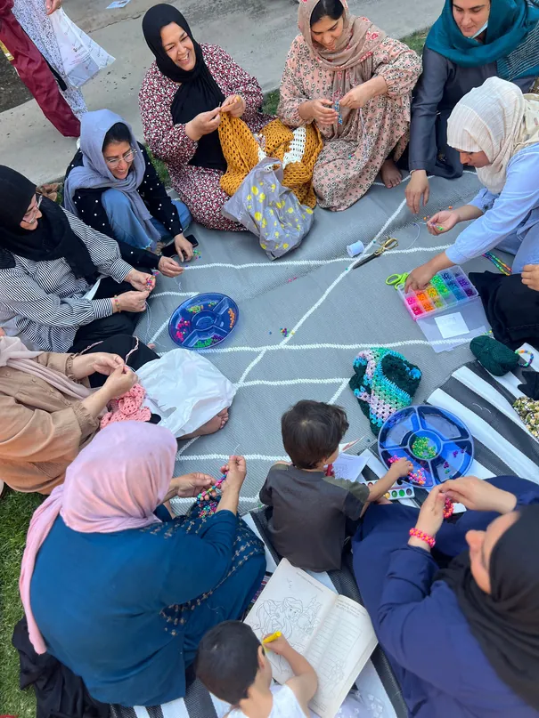 Women and children sitting on the floor in a circle at a FIRM craft workshop, working with yarn and fabric.